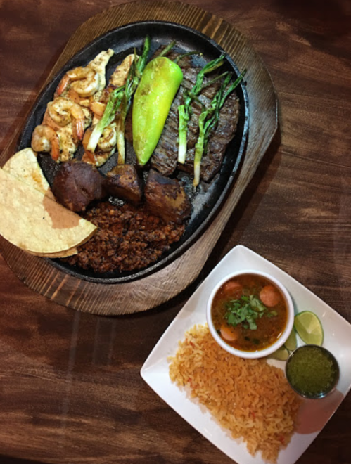 A sizzling platter with grilled shrimp, steak, and vegetables, served with rice, beans, and a side of salsa.