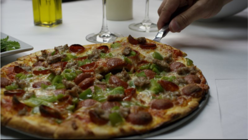 A hand using a knife to slice a pepperoni and sausage pizza topped with green peppers on a table.