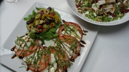 A plate with sliced tomatoes, mozzarella, and basil drizzled with balsamic, alongside a salad with olives and greens.