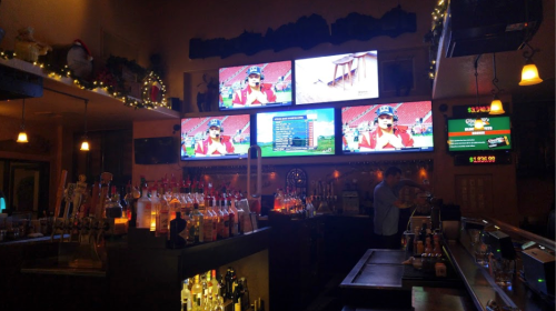 A dimly lit bar with multiple screens showing a sports event, shelves lined with bottles, and a bartender in the background.
