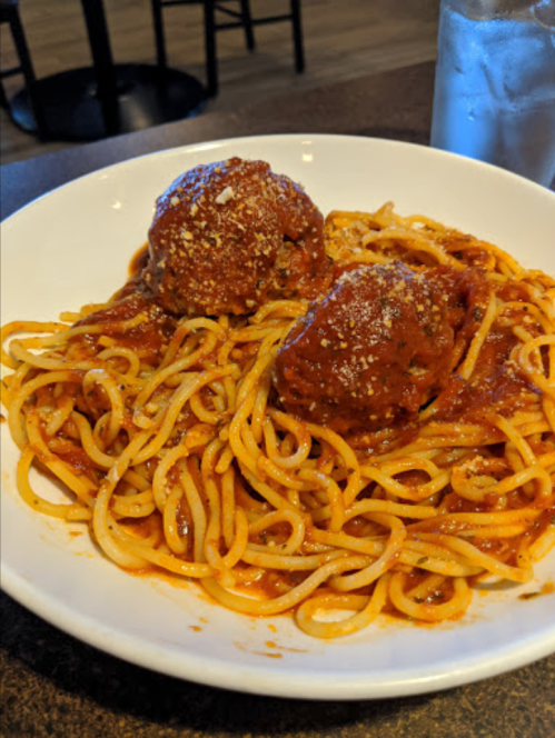 A plate of spaghetti topped with two meatballs and marinara sauce, served with a glass of iced water in the background.