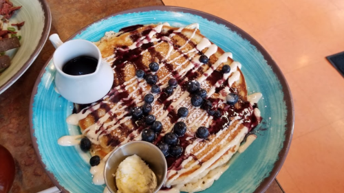 A large pancake topped with blueberries, drizzled with syrup and cream, served with a side of butter.