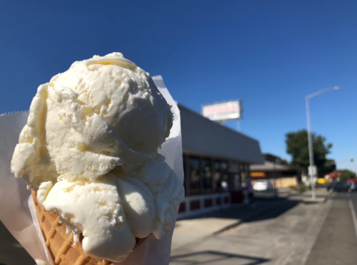 A close-up of a vanilla ice cream cone held in front of a shop on a sunny day.