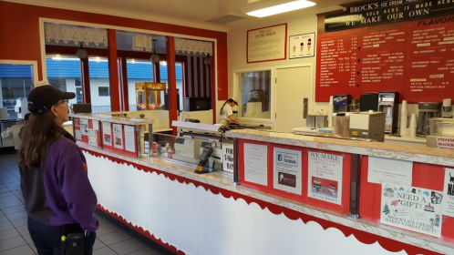 A woman stands in an ice cream shop, looking at the menu board while an employee prepares orders behind the counter.
