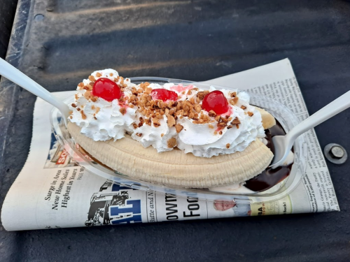 A banana split topped with whipped cream, cherries, and nuts, served on a newspaper.