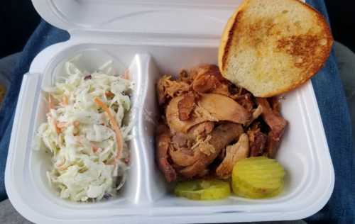 A takeout container with pulled pork, coleslaw, pickles, and a toasted bun on the side.