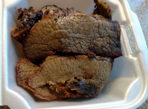 Sliced, cooked brisket in a foam takeout container, showing a smoky, charred exterior and tender texture.