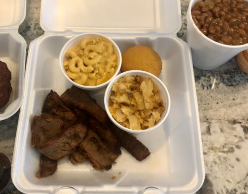 A takeout container with sliced brisket, macaroni and cheese, cornbread, and hash browns.