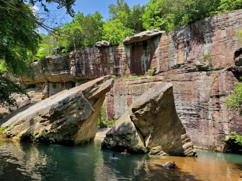 A serene natural scene featuring a rocky cliff beside a calm water pool, with people swimming in the water below.