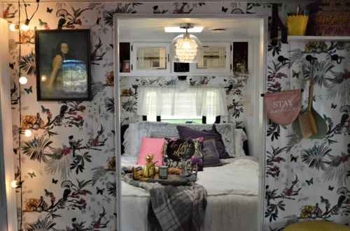 Cozy bedroom with floral wallpaper, a bed adorned with colorful pillows, and decorative items on a table.