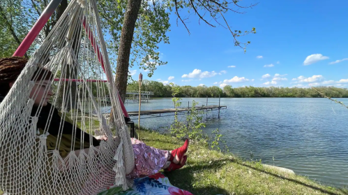 A person relaxes in a hammock by a serene lake, surrounded by trees and a clear blue sky.