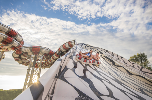 Two people slide down a large water slide under a blue sky with clouds, surrounded by colorful water park structures.