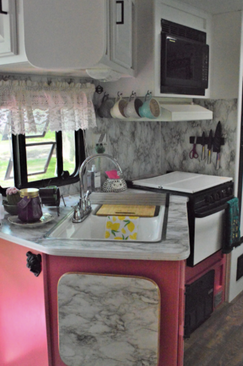 A cozy kitchen in an RV featuring a marble countertop, sink, stove, and colorful decor. Natural light filters in.
