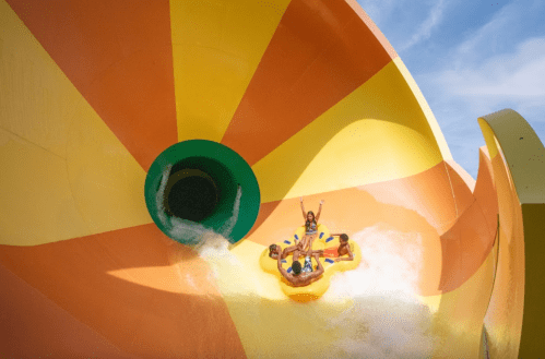 A group of four people on a colorful water slide, splashing down into a pool below on a sunny day.
