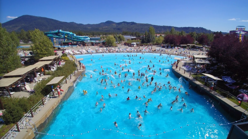 A busy water park with a large pool, water slides, and many people enjoying a sunny day. Mountains in the background.