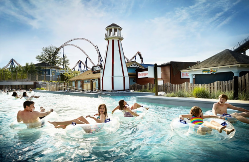 People float on inner tubes in a lazy river at a water park, with a lighthouse and roller coaster in the background.