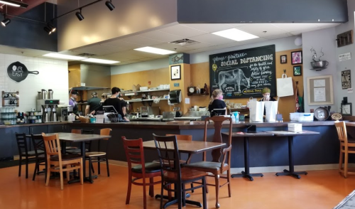 A cozy café interior with tables, a counter, and staff preparing food, emphasizing social distancing measures.