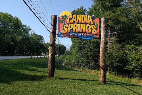 Sign for Candia Springs Adventure Park, featuring vibrant colors and tropical imagery, set against a green landscape.