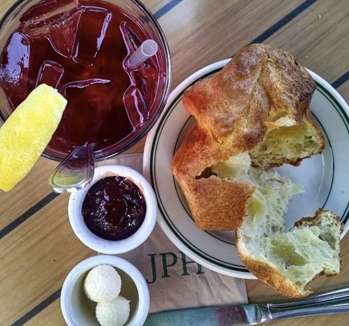 A plate with a split roll, jam, and a drink with ice and a lemon wedge, set on a wooden table.