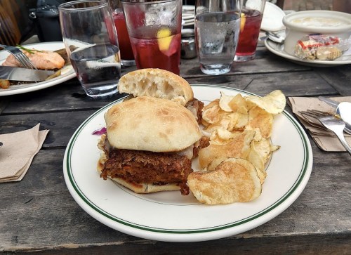 A sandwich with crispy fried chicken on a bun, served with potato chips and drinks in the background on a wooden table.