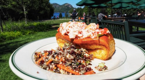 A lobster roll topped with lobster salad, served with a quinoa salad, set against a scenic outdoor backdrop.