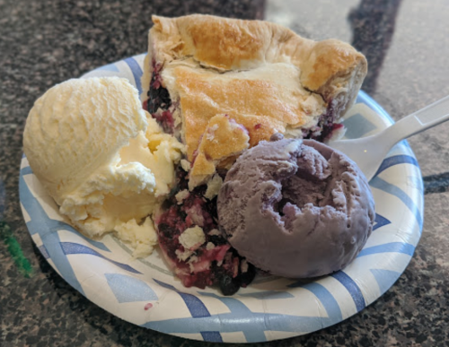 A slice of berry pie with a scoop of vanilla and a scoop of purple ice cream on a paper plate.
