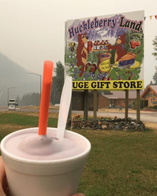 A hand holds a cup of huckleberry ice cream in front of a colorful Huckleberry Land sign. Smoky mountains in the background.