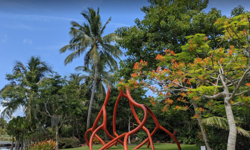 A vibrant outdoor scene featuring red abstract sculptures, lush green trees, and colorful flowers under a blue sky.