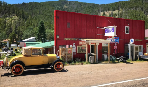 A vintage yellow car parked in front of a red general store surrounded by trees and mountains.