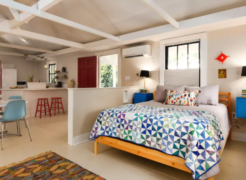 Cozy bedroom with a colorful quilt, modern decor, and an open layout featuring a dining area and kitchen in the background.
