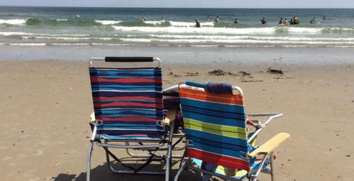 Two colorful beach chairs facing the ocean, with people swimming in the background and gentle waves lapping at the shore.