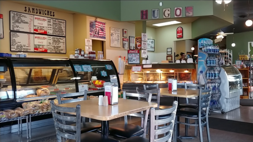 Interior of a casual deli with a menu board, display cases, and tables set for dining.