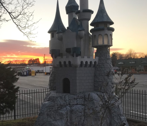 A whimsical castle structure with turrets against a vibrant sunset sky, surrounded by a fence and parking lot.