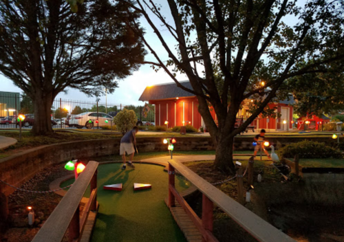 A mini-golf course at dusk, featuring colorful lights, trees, and a player preparing to putt.