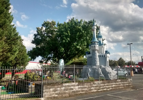 A whimsical castle structure surrounded by greenery and a fence, set against a blue sky with fluffy clouds.
