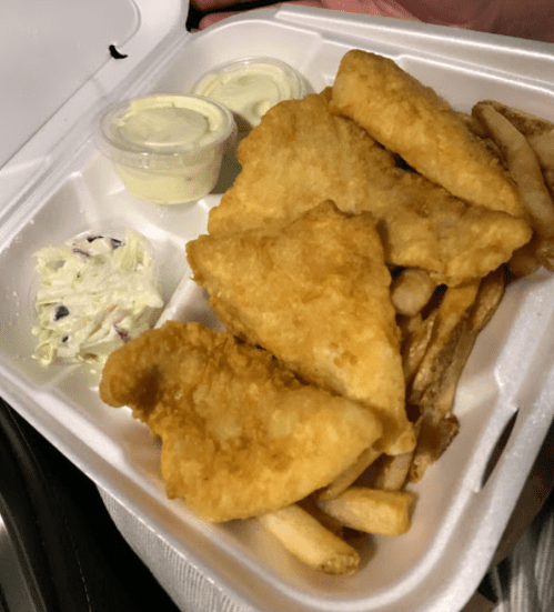 A takeout container with golden fried fish, fries, and two small cups of sauce, alongside a side of coleslaw.