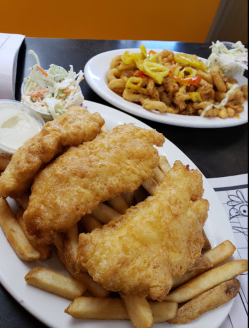 A plate of golden fried fish fillets on a bed of fries, with a side of coleslaw, and another plate of fried seafood in the background.