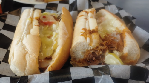 Two sandwiches on a black and white checkered paper, one with sliced meat and toppings, the other with pulled pork and pickles.