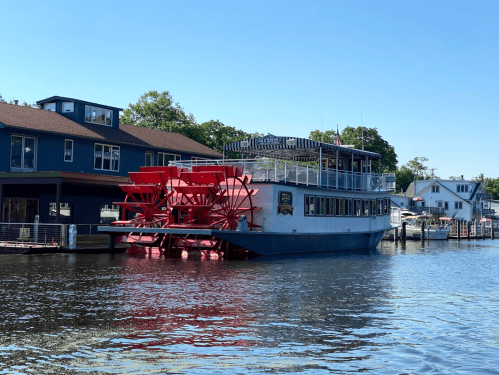 A large paddleboat with red wheels docked by a waterfront, surrounded by buildings and trees under a clear blue sky.