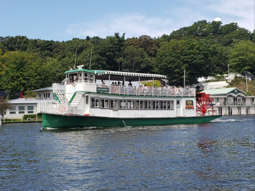 A green and white paddleboat with a red wheel sails on a calm lake, surrounded by lush greenery and buildings.