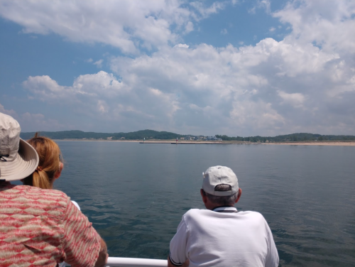 Two people on a boat gaze at a distant shoreline under a partly cloudy sky, reflecting on calm water.