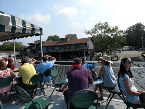 A group of people sitting on a boat, enjoying a scenic view of a riverside community on a sunny day.