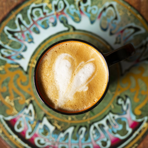 A close-up of a coffee cup with heart-shaped latte art, placed on a decorative, colorful plate.