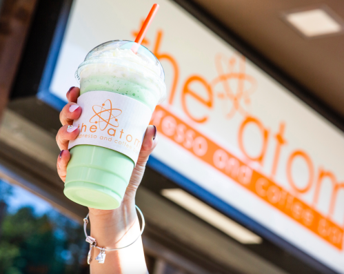 A hand holds a green beverage in a clear cup with a straw, in front of a café sign that reads "the atom."