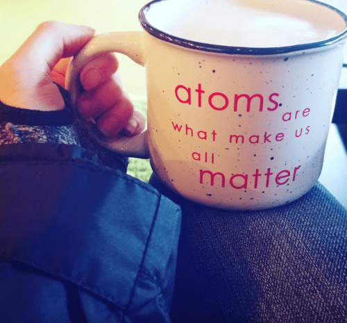 A hand holding a large mug with the text "atoms are what make us all matter" in pink on a speckled white background.