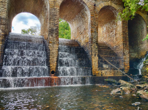 A stone structure with arches over cascading waterfalls, surrounded by greenery and reflecting water below.