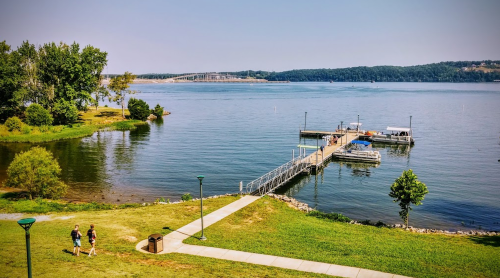 A serene lake view with a dock, surrounded by greenery and a walking path, under a clear blue sky.