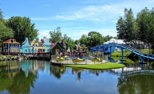 Colorful amusement park scene with rides and a pond, surrounded by trees under a clear blue sky.