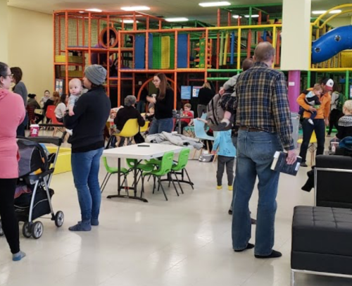 A busy indoor play area with families, children, and a playground structure, featuring people socializing and playing.