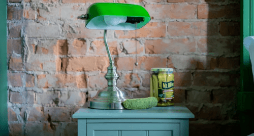 A green lamp and a jar of pickles sit on a small table against a brick wall. A pickle rests beside the jar.